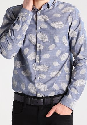 Homme portant une chemise bleue boutonnée avec des motifs de feuilles blanches, ceinture noire et pantalon noir, une main touchant le cou et l'autre main dans la poche.