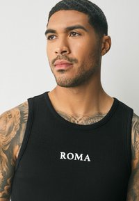 Sort ribbet tanktop med hvid "ROMA" print på forsiden. Designet har en rund halsudskæring og brede ærmegab. Glat tekstur.