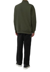 Olivgrüner Pullover mit hohem Kragen, lockerer Passform und geripptem Saum, kombiniert mit schwarzen Hosen und weißen Turnschuhen, von hinten gezeigt.