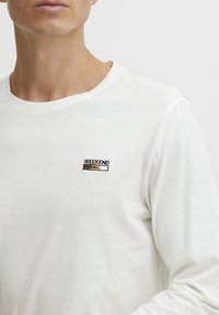 Hvid langærmet t-shirt lavet af blødt stof, prydet med et lille broderet "WEEKEND LOADING" logo i sort og gult på brystet.