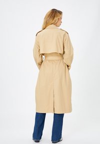 Beige trench coat dengan pinggang yang diikat, epaulet bahu, dan celah belakang, terbuat dari bahan yang halus dan ringan. Dipakai di atas denim.