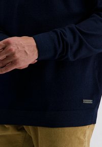 Navyblauer Strickpullover mit gerippten Ärmelbündchen und Saum, versehen mit einem metallenen Logoschild an der unteren Seite. Getragen über beigen Cordhosen.