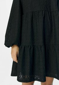 Robe noire à volants avec des manches longues, présentant un tissu texturé avec un motif en diamant. L'ourlet se situe au-dessus du genou, laissant passer la lumière à travers le matériau.