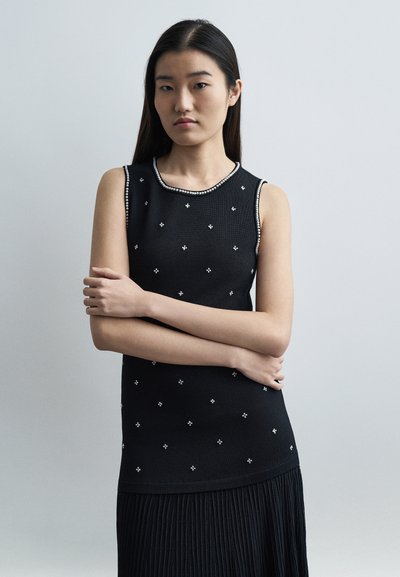 Jeune femme aux longs cheveux bruns portant un haut noir sans manches avec de petits motifs floraux blancs et une jupe plissée, debout les bras croisés.