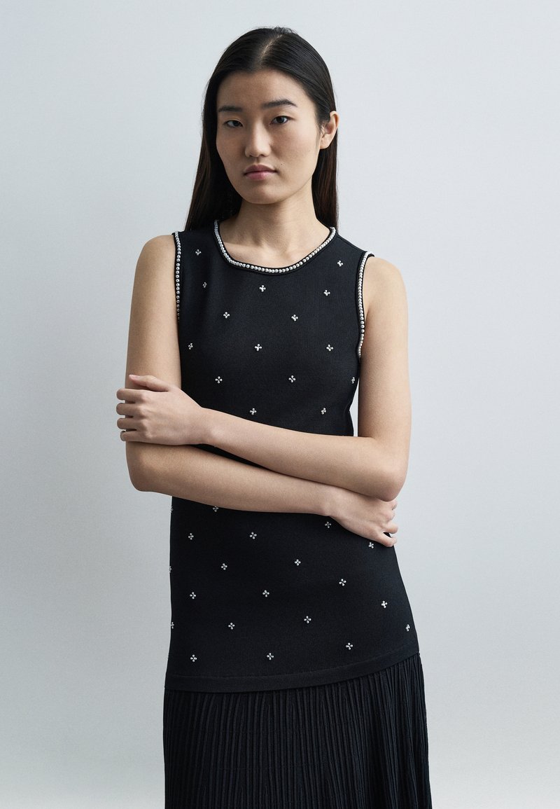 Νέα γυναίκα με μακριά σκούρα μαλλιά φοράει αμάνικο μαύρο τοπ με μικρά λευκά λουλουδάτα σχέδια και πλισέ φούστα, στέκεται με σταυρωμένα χέρια.