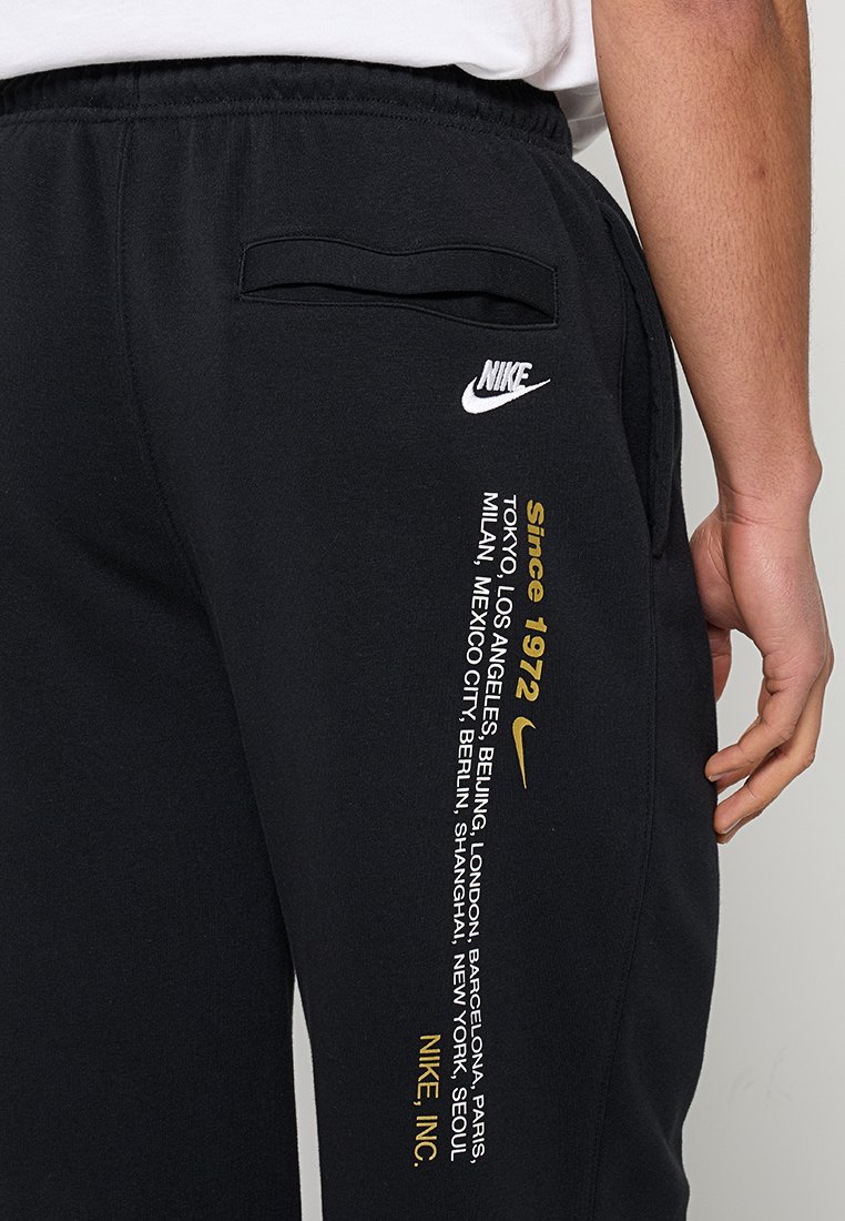 Pantalon de survêtement Nike noir avec texte blanc et or listant des villes et "Depuis 1972" sur la cuisse droite, porté avec un t-shirt blanc.