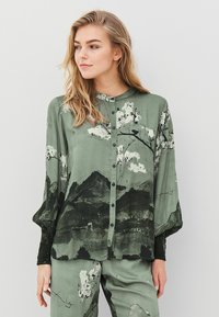 Grøn, silkebluse med knaplukning foran, prydet med et bjerg- og blomsterprint i sort og hvidt. Lange ærmer med strukturerede sorte manchetter.