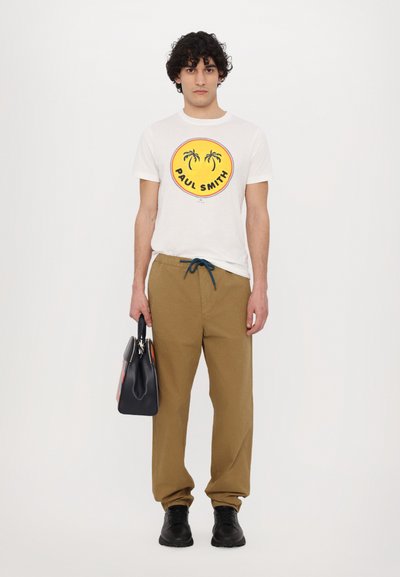 White t-shirt with a yellow graphic featuring palm trees and "PAUL SMITH." Brown trousers with a drawstring, holding a black bag. Black shoes.