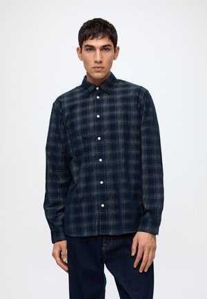Jeune homme portant une chemise à carreaux bleu foncé à manches longues boutonnée et un jean foncé sur un fond clair uni.