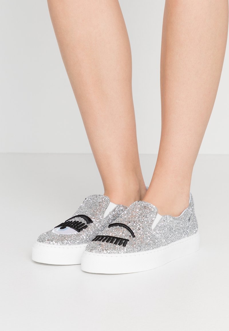 Ben som bär glittriga silverfärgade slip-on sneakers med svart ögonfrans- och ögonbroderi på vita platåsålar mot en enkel bakgrund.