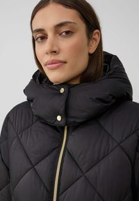 Veste matelassée noire avec un col haut en style col cheminée, dotée d'une fermeture éclair dorée et de détails à boutons-pression. Texture lisse et motif en losanges.