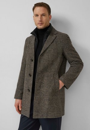 Mann trägt einen grauen karrierten Mantel über einer schwarzen Reißverschlussjacke und einem schwarzen Rollkragenpullover, steht vor einer hellgrauen Wand.