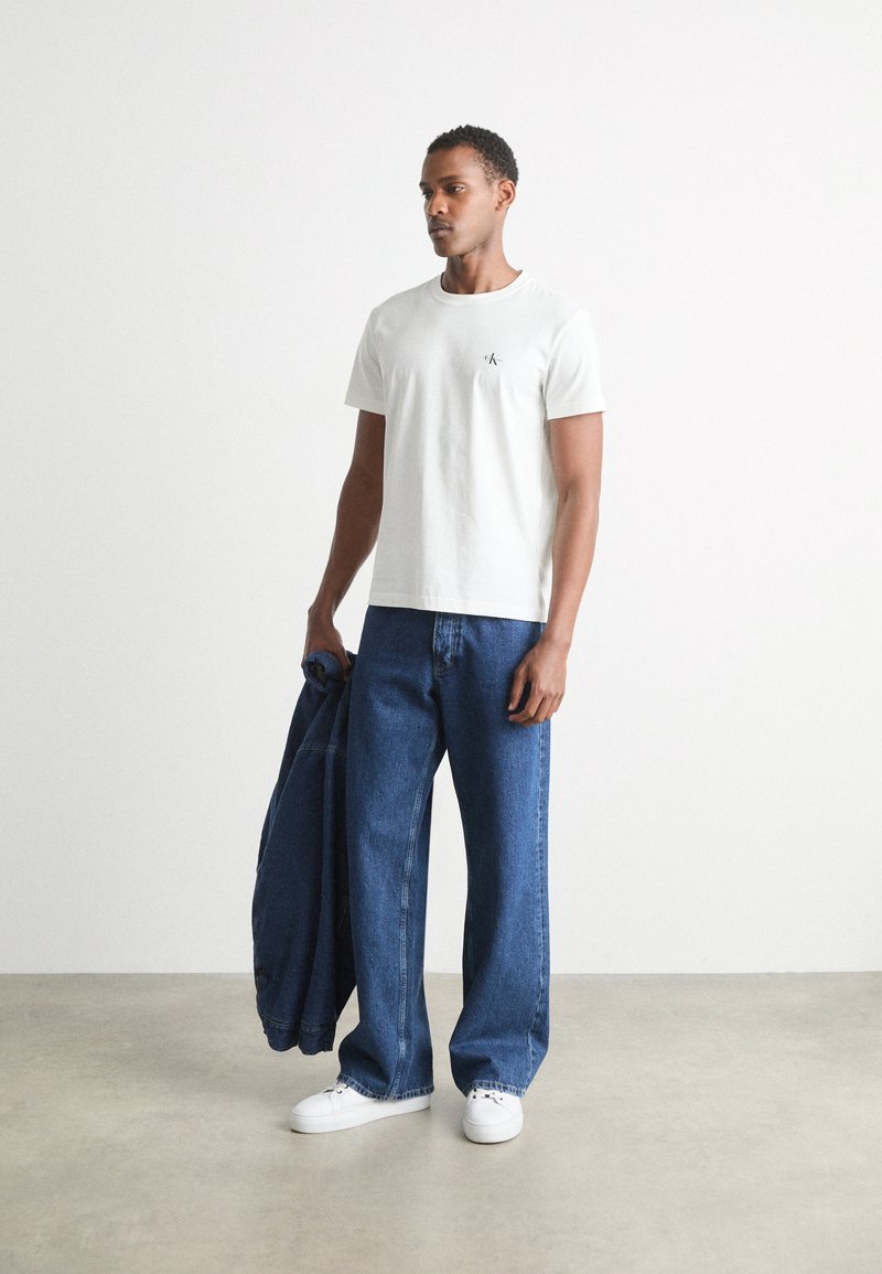 Hvid bomulds T-shirt med et lille logo, parret med løse blå denimjeans. Modellen holder en mørk denimjakke. Hvide sneakers fuldender outfittet.