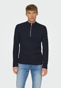 Marinefarbener Grobstrickpullover mit hoch geschlossenem Kragen und halbem Reißverschluss. Mit gerippten Bündchen und Saum, aus einem strukturierten Stoff gefertigt. Kombiniert mit blauen Jeans.