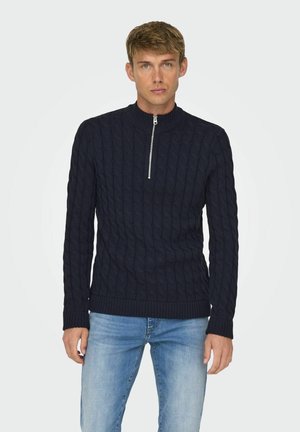 Marinblå kabelstickad tröja med hög krage och halv dragkedja. Ribbade muddar och nederkant, tillverkad av ett texturerat tyg. Kombineras med blå jeans.