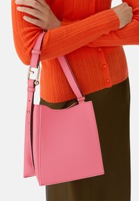 Roze leren handtas met een gestructureerd oppervlak, voorzien van een gebogen bovenkant, verstelbare schouderband en gouden slot. Gedragen over een oranje trui.