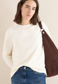 Cream-colored long-sleeve top with a round neckline and subtle seam detail, paired with dark denim jeans and a brown suede shoulder bag.