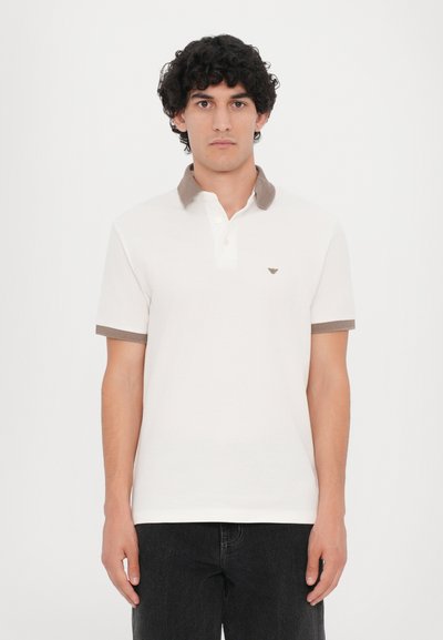 Polo shirt blanc avec un col et des manches côtelés marron. Présente un petit logo sur la poitrine gauche. Fabriqué en coton avec une texture lisse.