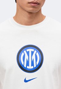Gros plan d’une personne portant une chemise blanche avec le logo bleu et noir de l’Inter Milan et un swoosh Nike bleu en dessous.