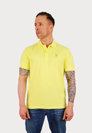 Yellow polo shirt with short sleeves and a classic collar. Features a small embroidered logo on the chest. Worn with blue jeans.