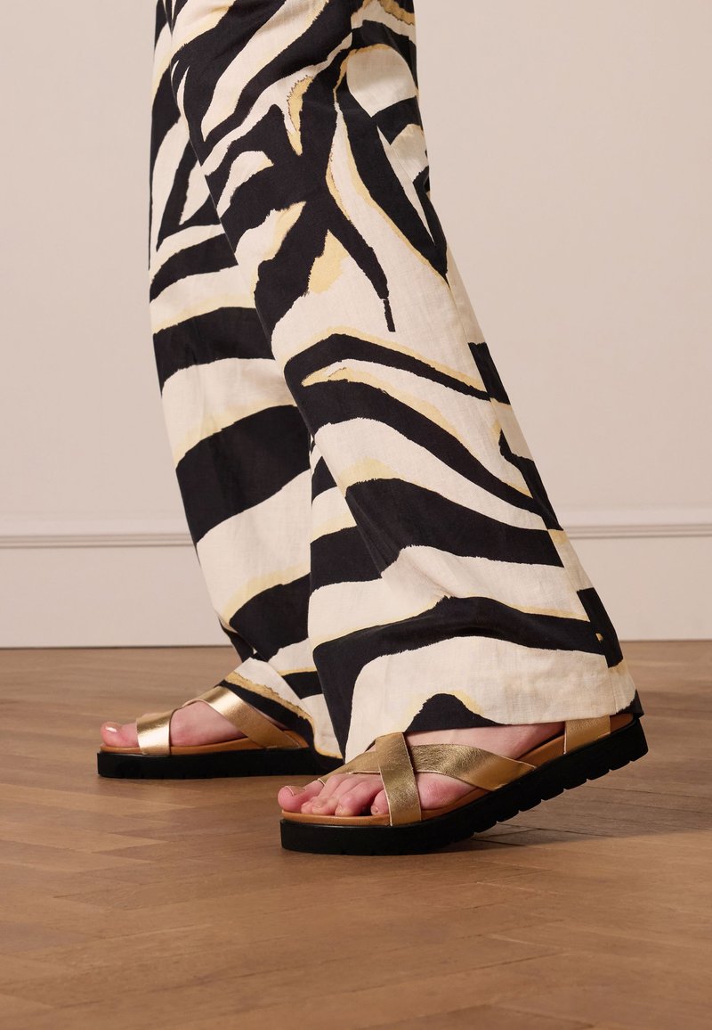 Gouden metallic sandalen met een platte zwarte zool, gecombineerd met zwart-witte zebra-streeppantalons met gouden accenten en een wijd uitlopende pasvorm.
