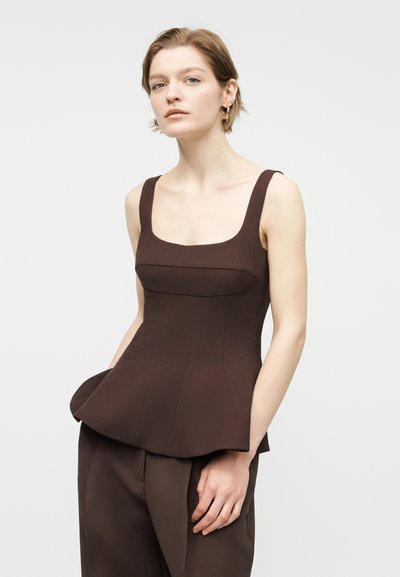 Γυναίκα με αμάνικο εφαρμοστό τοπ σε σκούρο καφέ χρώμα με τετράγωνη λαιμόκοψη και αντίστοιχο παντελόνι, στέκεται με το ένα χέρι στην τσέπη, μπροστά σε απλό φόντο.