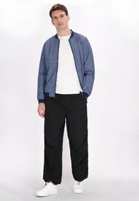 Blaue gemusterte Bomberjacke mit gerippten Bündchen und marineblauem Kragen, kombiniert mit lockeren schwarzen Hosen und weißen Sneakers. Einfaches, modernes Design.