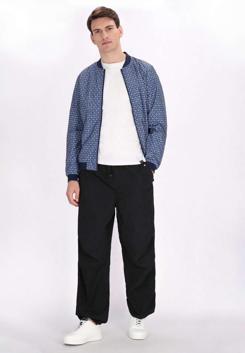 Blaue gemusterte Bomberjacke mit gerippten Bündchen und marineblauem Kragen, kombiniert mit lockeren schwarzen Hosen und weißen Sneakers. Einfaches, modernes Design.