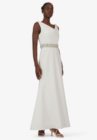 Weißes, bodenlanges Kleid mit einem Wasserfallausschnitt und einem mit Perlen verzierten silbernen Gürtel. Glatte Stoffoberfläche mit einer fließenden Silhouette und minimaler Detailgestaltung.