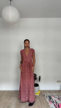 Frau mit geflochtenem Haar, die ein rotes gemustertes Maxikleid und schwarze Stiefel trägt, steht in einem minimalistischen Zimmer mit einer Pflanze und einer hängenden weißen Laterne.