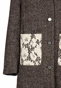 Brun fiskbensmönstrad kappa med svarta knappar och stora fickor fram i vitt blommigt spetsmaterial.