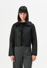 Femme portant une veste matelassée noire courte avec des liens, associée à un pantalon gris texturé taille haute, se tenant devant un fond blanc.