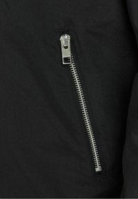 Tissu de veste noire avec une fermeture éclair métallique, présentant un design distinctif à dents blanches et une finition élégante. Fermeture éclair positionnée en angle.