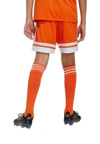 Orangene Fußballuniform mit Shorts mit weißen Streifen und passenden knielangen Socken. Schwarze Fußballschuhe mit strukturierten Sohlen.