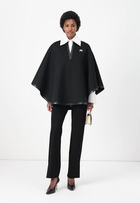 Juodas pončas su užtrauktuku, po juo balta marškinėlių apykakle, derinamas su juodomis kelnėmis ir aukštakulniais. Lygi tekstūra, modernus dizainas.