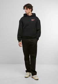 Schwarzer Hoodie mit einer Fronttasche und Kapuze mit Kordelzug, mit rotem besticktem Logo. Kombiniert mit schwarzen Hosen und beigen Sneakern.