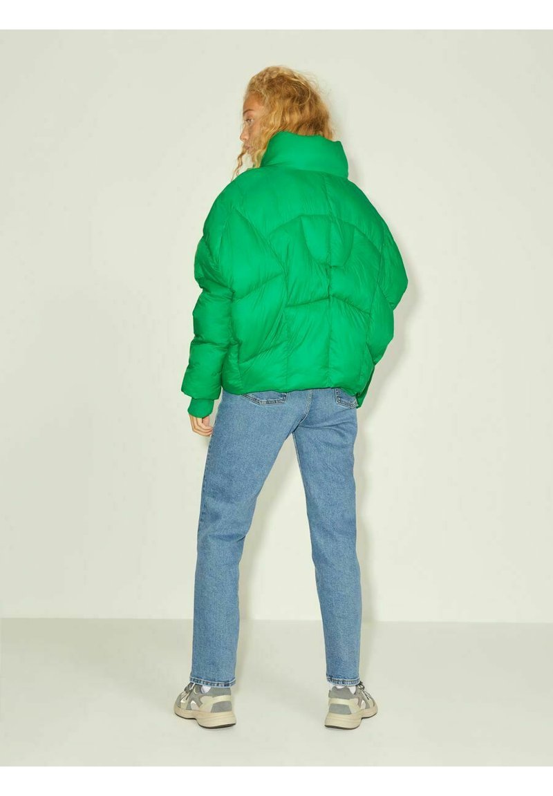 Giacca a vento verde con colletto alto, texture trapuntata e vestibilità over-size, abbinata a jeans azzurri chiari e sneakers grigie.