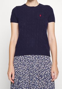 Žena v námořnickém modrém krátkém svetru s pleteným vzorem a malým červeným logem na kapse, doplněném o květovanou sukni v námořnické barvě.