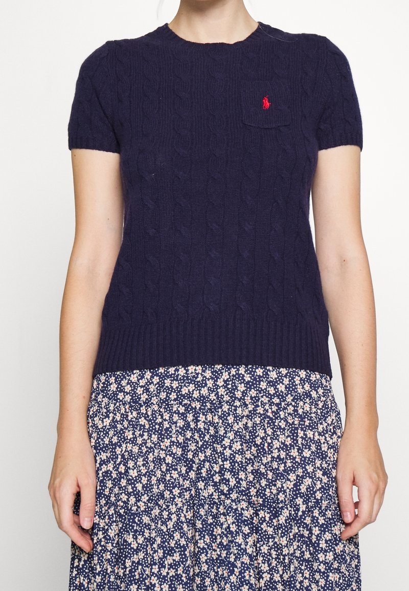 Žena v námořnickém modrém krátkém svetru s pleteným vzorem a malým červeným logem na kapse, doplněném o květovanou sukni v námořnické barvě.