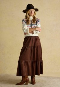 Pletený krémový sveter s farebnými geometrickými vzormi, skombinovaný s hnedou maxi sukňou s volánmi a hnedými čižmami na podpätku. Široký čierny klobúk.