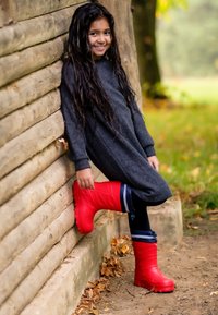 Rode rubberen laarzen met een gestructureerd oppervlak en gevormd patroon, gecombineerd met een donkergrijze geribbelde jurk, leunend tegen een houten muur.