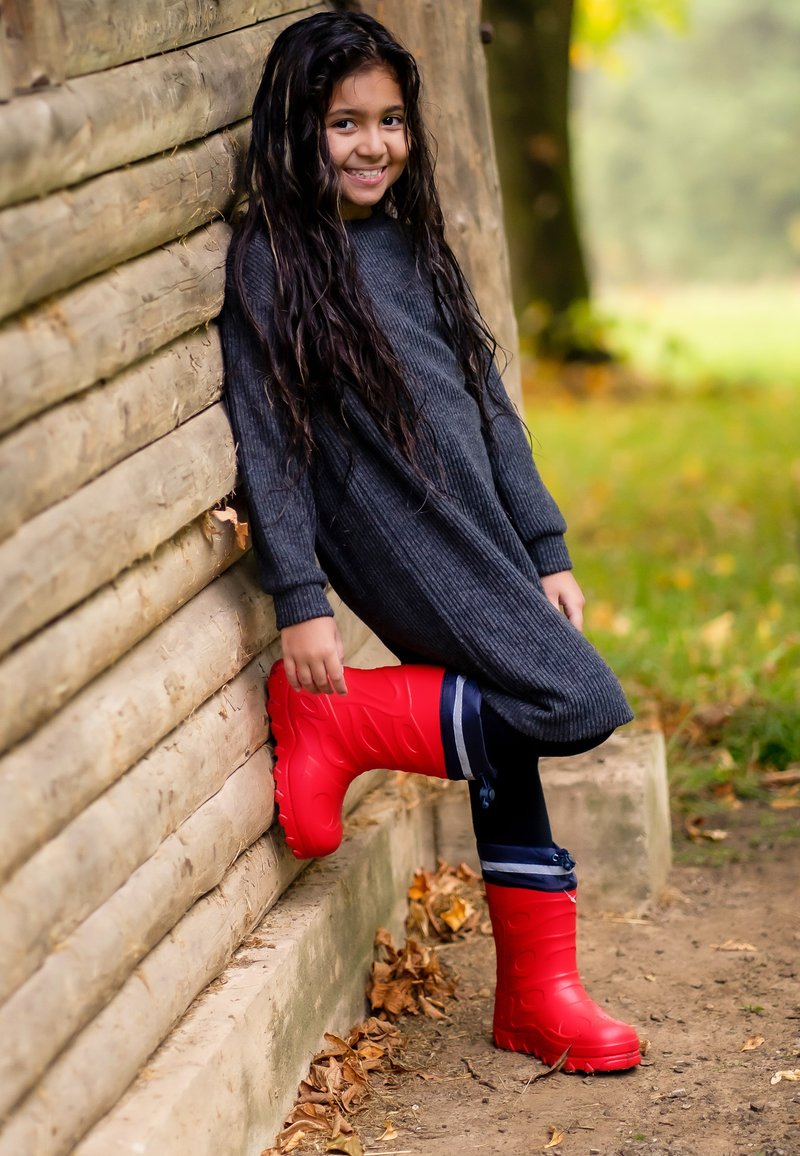 Rode rubberen laarzen met een gestructureerd oppervlak en gevormd patroon, gecombineerd met een donkergrijze geribbelde jurk, leunend tegen een houten muur.