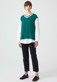 Gilet a coste verde con scollatura a V a strisce bianche e nere, indossato sopra una camicia bianca, abbinato a jeans neri a gamba dritta e mocassini.