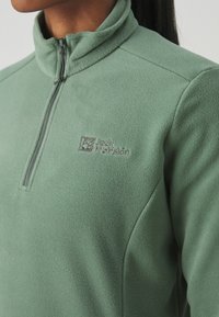 Groene fleece pullover met een hoge kraag, voorzien van een rits-halslijn en een geborduurd logo in grijs op het linker borstgebied.