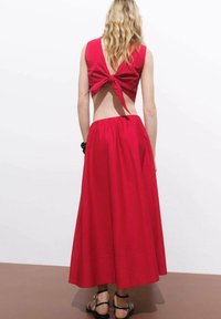 Abito lungo in cotone rosso con design senza maniche, una grande apertura sulla schiena e un fiocco legato sul retro. Presenta una silhouette ampia e fluida.