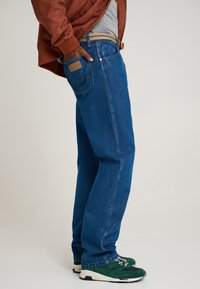 Blugi din denim albastru cu croială dreaptă, cusături vizibile și o etichetă de piele pe buzunarul din spate. Asortați cu pantofi sport verzi.