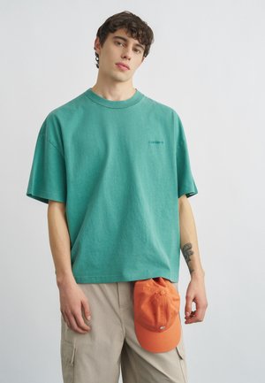 Joven con una camiseta holgada verde azulado y pantalones cargo beige, sosteniendo una gorra de béisbol naranja en su mano derecha.