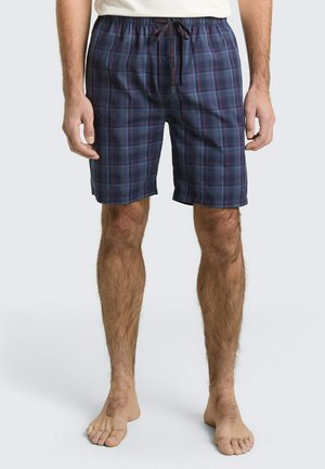 Homme portant un short écossais bleu marine et bordeaux jusqu'aux genoux avec une taille à cordon, debout pieds nus devant un fond uni.