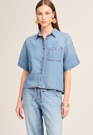 Young woman wearing loose-fitting short-sleeve denim shirt with front pocket and light blue denim jeans, accessorized with gold earrings and bracelets.
