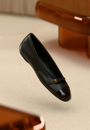 Scarpa piatta in pelle nera con puntale lucido nero, cinturino centrale con bottone dorato e superficie liscia.